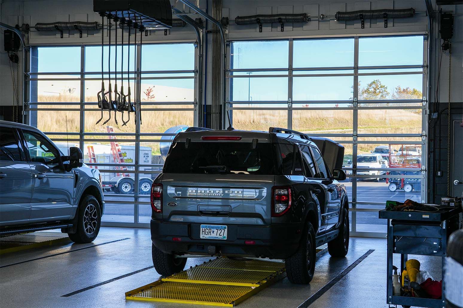 Interior service bay with a Ford Bronco Sport and a pickup on drive-on ramps in front of closed full-vision doors