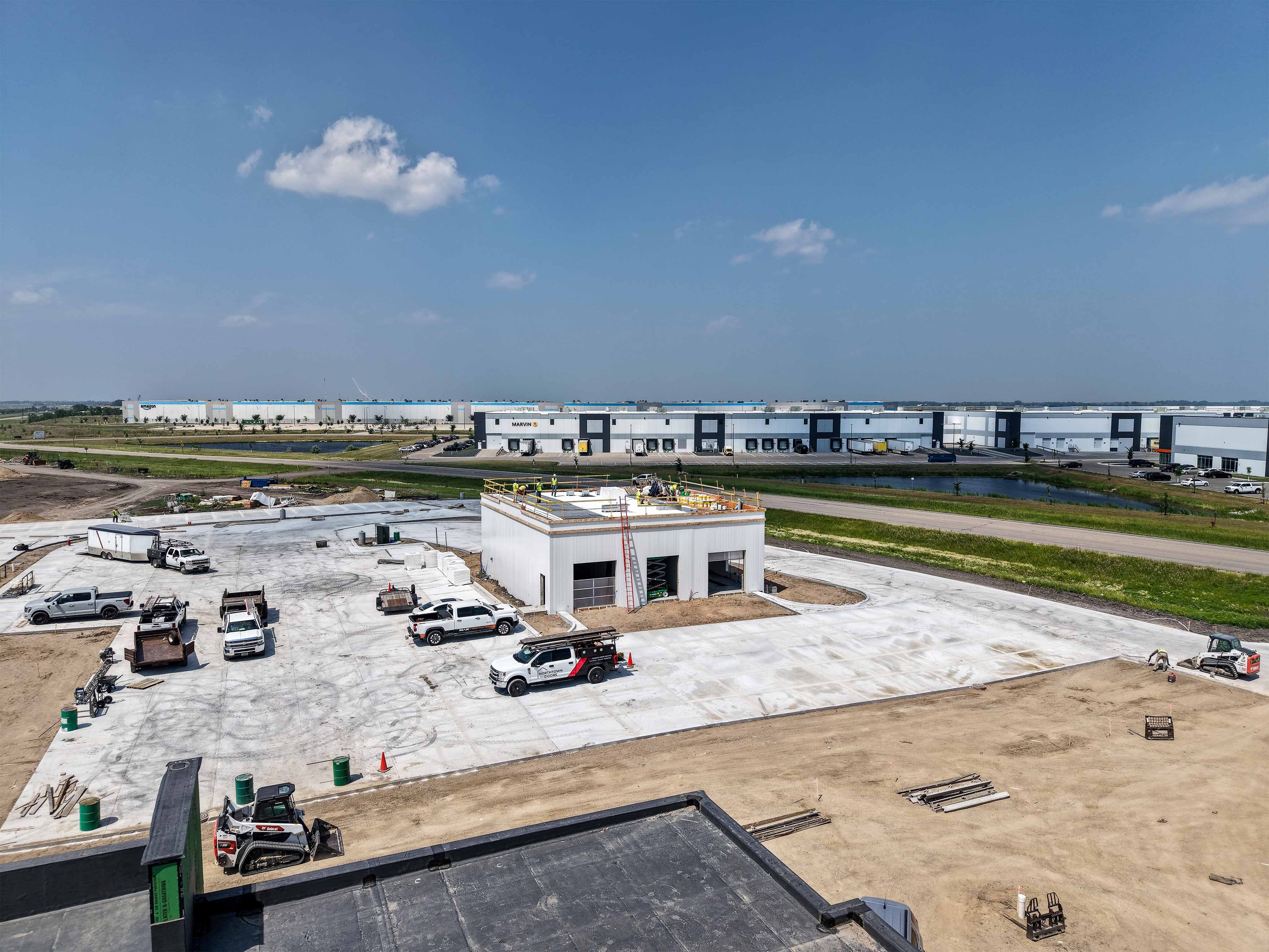 Wide aerial of the Northdale Oil travel center under construction beside the Interstate 29 corridor, with the Marvin facility and Amazon fulfillment center behind it